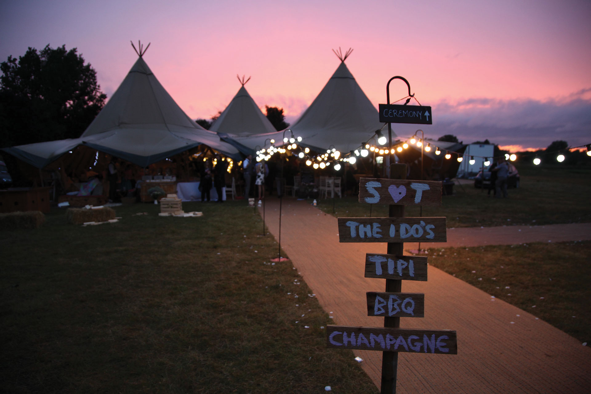 Wedding ceremony sign tipi/tents people and lights in background sunset