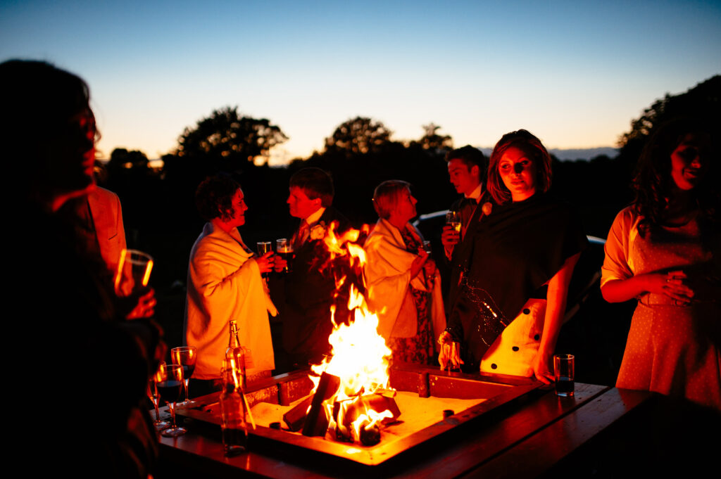 Firepit at a wedding with people standing around with drinks in the evening